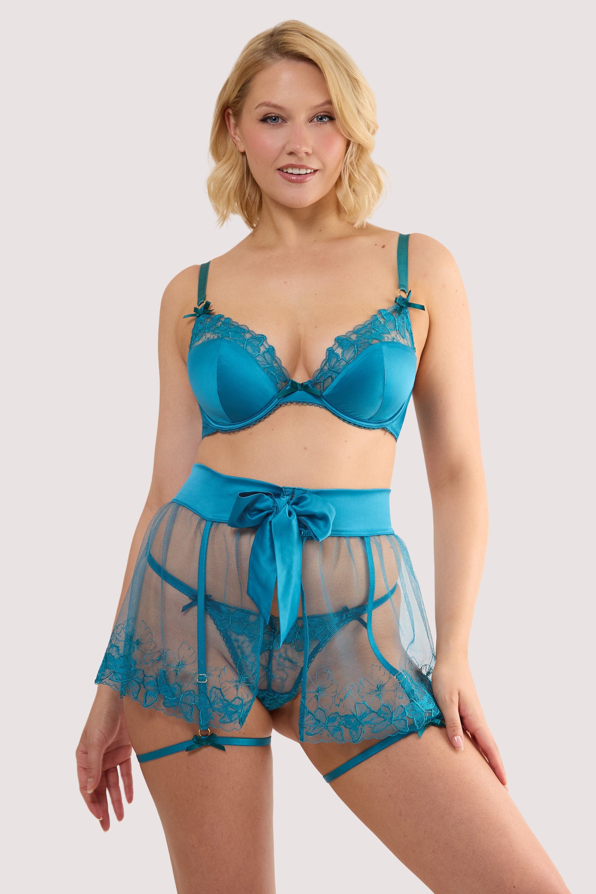 Model wears a mariposa teal butterfly embroidery skirted suspender with sheer mesh with teal butterfly embroidery, skirted suspender silhouette with a matching thong and bra.
