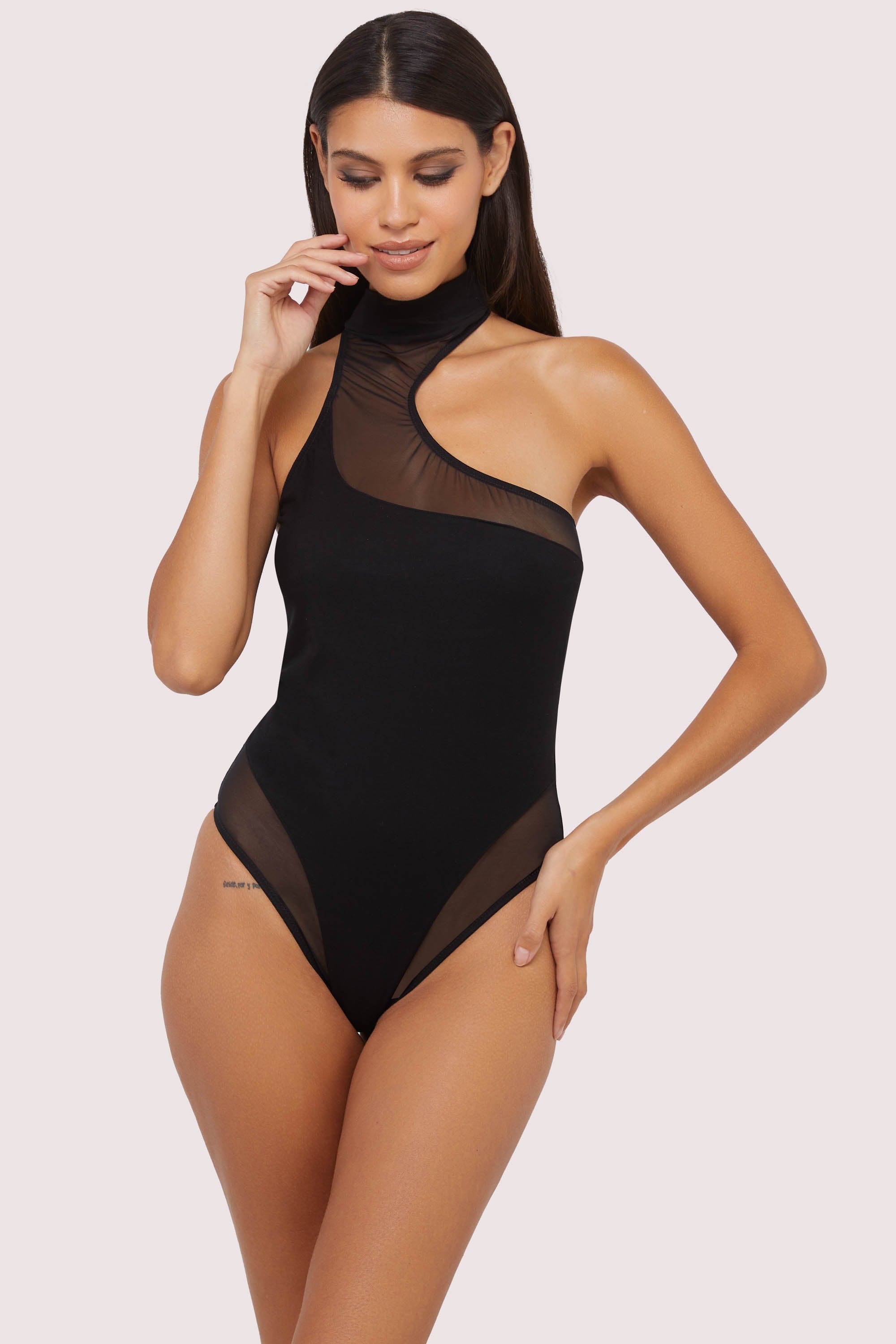 Model wearing black asymmetric mesh and jersey thong bodysuit with high neck and sheer panel detailing